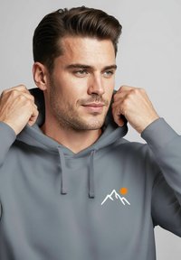Grauer Hoodie aus weichem Material, mit einer vorderen Kängurutasche und einem Berggrafik mit einer orangefarbenen Sonne. Mit einem Kordelzugkapuze und gerippten Bündchen.