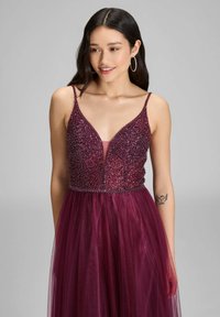 Burgundowa suknia wieczorowa z koralikowym dekoltem, głębokim V i przezroczystą tiulową spódnicą. Posiada cienkie ramiączka i zwiewny fason.