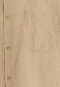 Camisa de lino beige con cuello doblado, cierre frontal de botones y un bolsillo en el pecho de forma angular. Tejido suave y texturizado.