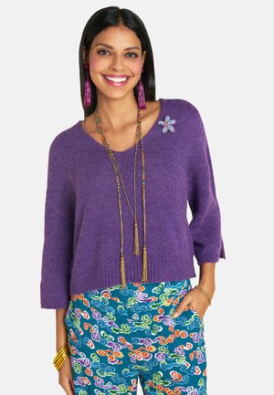 Lila gestrickter Pullover mit 3/4-Ärmeln, versehen mit einer floralen Brosche. Kombiniert mit mehrfarbigen, gemusterten Hosen. Goldene Schmuckakzente sind inklusive.