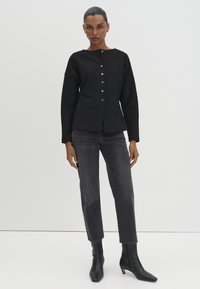 Woman wearing black buttoned long-sleeve top, dark gray straight-leg jeans, and black ankle boots, standing against a plain light background.