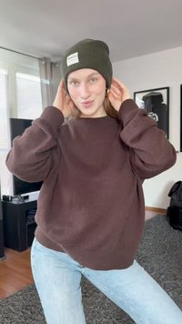 Maglione marrone a coste con maniche oversize, abbinato a un berretto di lana verde scuro con un'etichetta bianca. Jeans azzurri chiari. Ambiente interno.