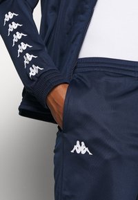 Personne portant un survêtement bleu marine Kappa avec des logos blancs imprimés sur la manche et la poche du pantalon, main partiellement dans la poche.