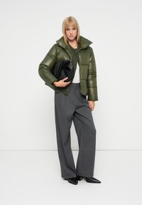 Moteris, vilkinti alyvuogių žalia pūkine striuke, tamsiai pilkomis plačiomis kelnėmis ir juodais smailiais bateliais, laiko sulankstytą juodą odinį maišelį.