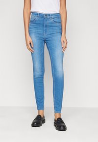 Persona vestida con jeans ajustados azul claro y mocasines negros, de pie contra un fondo blanco liso.