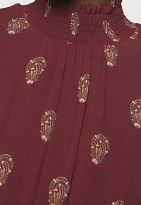 Tissu bordeaux avec petits motifs floraux paisley beige et vert, doté d'un col montant froncé et volanté.