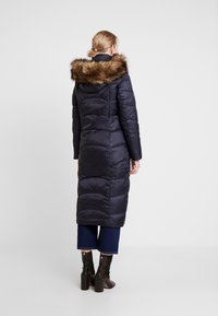 Dlouhý námořnicky modrý prošívaný kabát s kapucí lemovanou kožešinou, s obdélníkovými prošitými sekcemi a hladkou strukturou. Kombinován s tmavými džínami a botami.