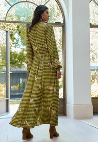 Gara, ķīļveida kleita dzeltenā un zaļā krāsā ar ziedu akcentiem; ir garas piedurknes un pieguļošs jostasvieta. Nēsāta ar brūnām papēžu zābakiem.