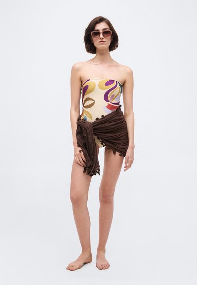 Γυναίκα με ασύμμετρο ολόσωμο μαγιό με λουλούδια και καφέ φούστα τύπου wrap, με κοντά μαλλιά και γυαλιά ηλίου, στεκόμενη ξυπόλυτη μπροστά σε απλό φόντο.