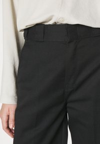 Gros plan d'une personne portant un pantalon noir taille haute et une chemise crème à manches longues, avec une main visible à côté de la hanche.