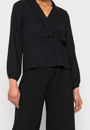 Vrouw draagt een zwarte lange mouw V-hals blouse met geplooide taille en bijpassende zwarte wijde broek, afgebeeld vanaf midden torso.