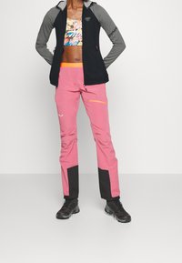 Giacca nera e grigia con zip e maniche lunghe, abbinata a pantaloni rosa con dettagli neri e una tasca con zip, indossata con scarpe da escursionismo nere.