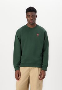 Green cotton sweatshirt with a ribbed crew neckline, long sleeves, and a small red embroidered logo on the chest. Paired with beige pants.