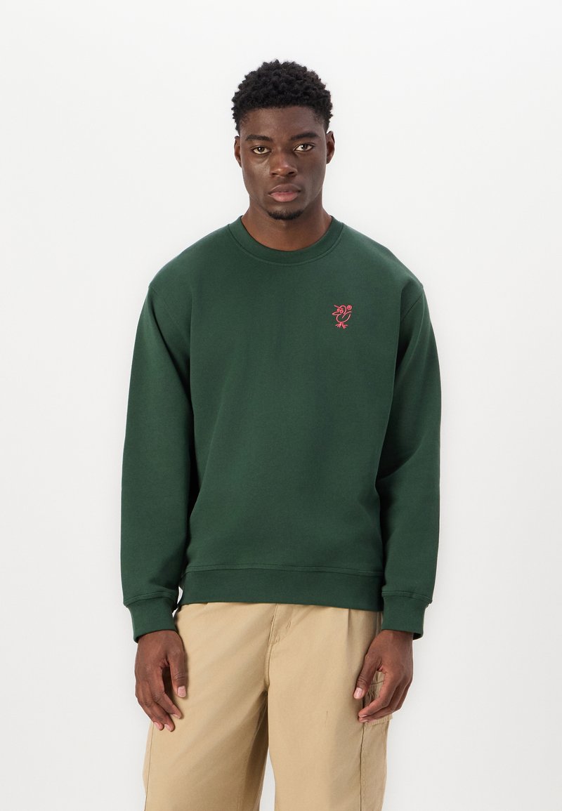 Green cotton sweatshirt with a ribbed crew neckline, long sleeves, and a small red embroidered logo on the chest. Paired with beige pants.