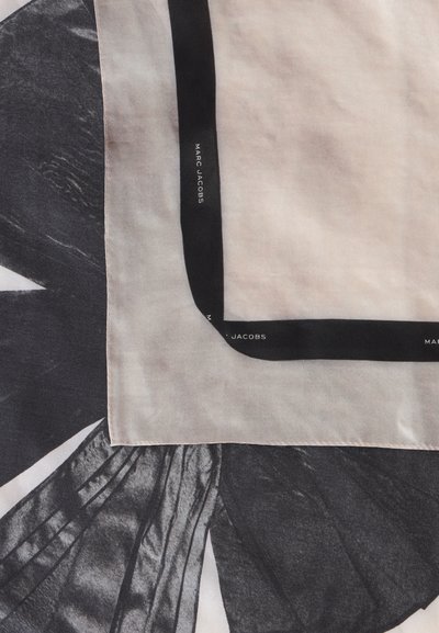 Écharpe beige clair avec une bordure noire portant le texte "MARC JACOBS", posée sur un fond de tissu texturé noir et blanc.