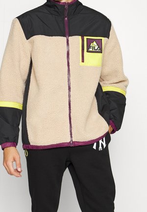 Beżowo-czarna kurtka polarowa z żółtymi akcentami, fioletowymi zamkami, naszywką z logo FILA na piersi, noszona przez osobę z ręką w kieszeni.