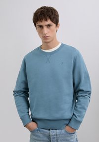 Joven con cabello corto castaño, vistiendo un suéter de cuello redondo azul claro y jeans de lavado claro, de pie con las manos en los bolsillos.