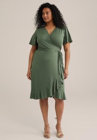 Rochie verde din material moale, cu mâneci scurte în volane, detalii cu volane pe lateral și talie legată. Materialul pare a fi moale și are o textură netedă.