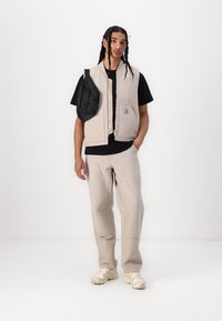 Beige polstret vest med to frontlommer, glidelås og logo-patch. Kombinert med svart t-skjorte og lysegrå bukser. Fottøyet er kremfarget.