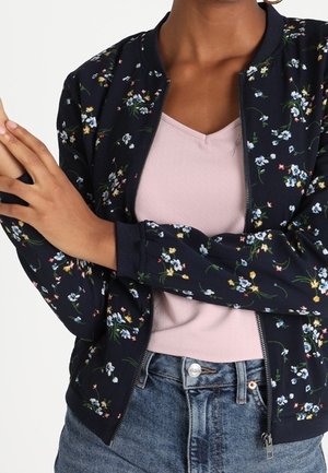 Vrouw draagt een marineblauw bloemenritsjack over een lichtroze V-hals top en een blauwe spijkerbroek, met gekruiste armen voor zich.