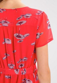 Červené šaty s krátkými rukávy, zdobené vzorem malých modro-bílých deštníků, stažené v pase odpovídajícím páskem.