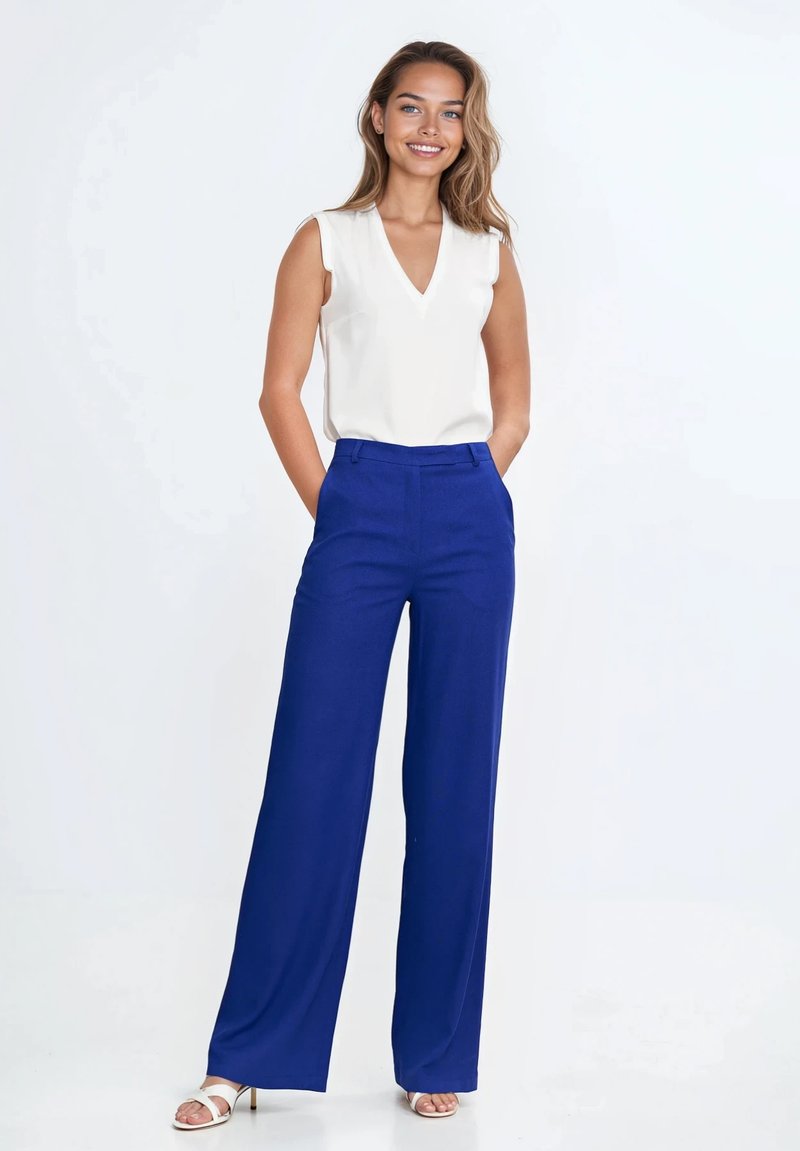 Biała, bezrękawnikowa bluzka z dekoltem w kształcie litery V, zestawiona z szerokimi, królewskoniebieskimi spodniami. Buty są białe, na cienkim obcasie. Gładkie tekstury w całym zestawie.
