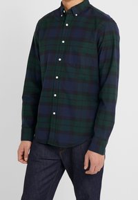 Camicia button-up in tartan verde scuro e blu navy. Presenta una tasca sul petto, bottoni bianchi e maniche lunghe. Il tessuto appare liscio e leggero.