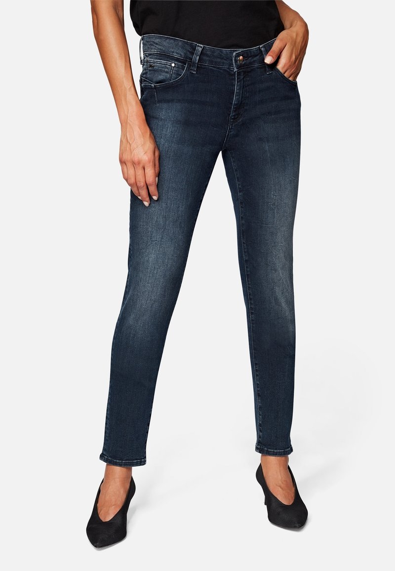 Femme portant un jean skinny bleu foncé et des talons aiguilles noirs, debout avec une main dans la poche et l'autre détendue le long du corps.