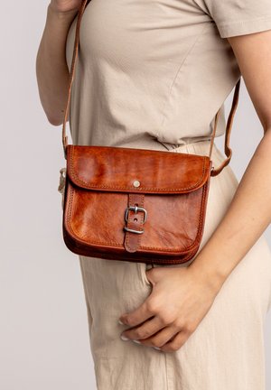 Frau in beiger Kleidung mit einer kleinen braunen Lederumhängetasche, die über einen Schnallenverschluss und eine Klappe verfügt.