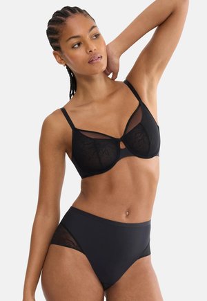 Femeie cu părul împletit, prezentând un sutien negru transparent din dantelă florală și chiloți asortați cu talie înaltă, pozând cu un braț ridicat pe fundal alb.