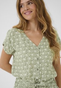 Blouse verte à boutons avec un motif abstrait blanc, manches courtes et décolleté en V. Style avec une taille nouée et un tissu léger et texturé.