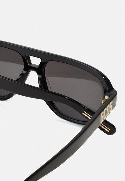 Black sunglasses with dark lenses, bold rectangular shape, and a cutout bridge design. Features gold accents and "GCDS" branding on the temple.