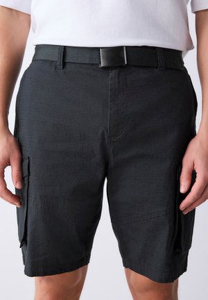 Hombre con pantalones cortos cargo negros con múltiples bolsillos y cinturón negro, combinado con una camisa blanca metida por dentro.