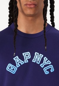 Navy blue t-shirt featuring arched text "GAP NYC" in light blue. The fabric appears smooth with a crew neckline.
