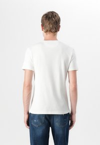 Hvit bomull t-skjorte med korte ermer, rund hals og et enkelt design. Kledd med blå jeans som har baklommer og subtil slitasje.