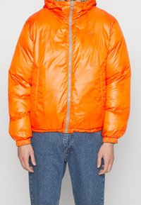 Personne portant une doudoune orange vif avec une fermeture éclair à l'avant et un jean bleu, debout contre un fond clair uni.