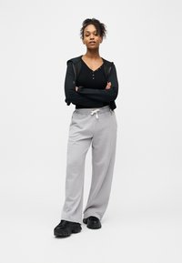 Jeune femme debout avec confiance, les bras croisés, portant un haut court noir, un sweat à capuche noir, un pantalon de survêtement gris et des baskets épaisses noires.