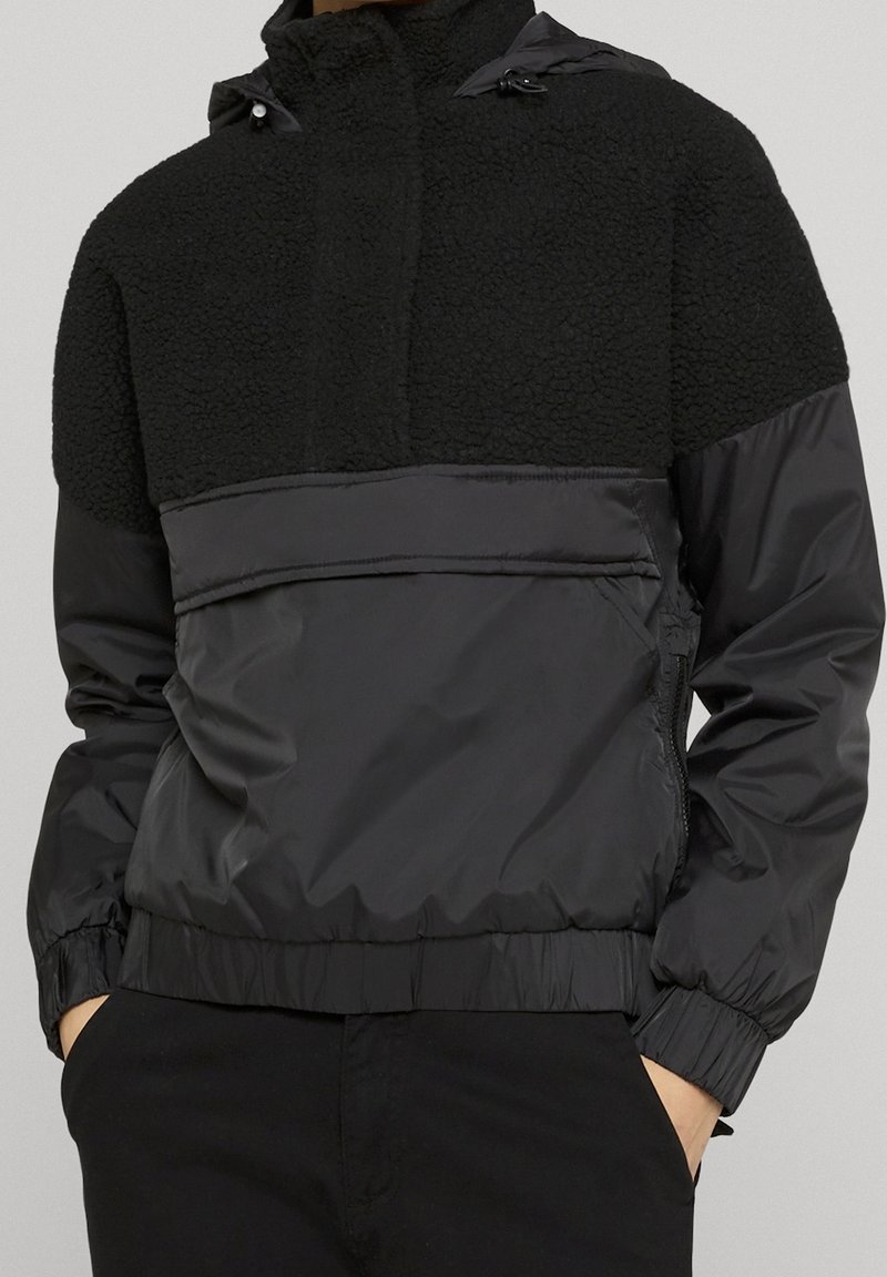 Homme portant un pull-over en polaire et nylon noir avec poche avant et poignets élastiques, mains dans les poches du pantalon.
