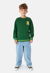 Grön sweatshirt med en gul "R"-logotyp, med randiga muddar och nederkant; ihop med ljusblå baggy jeans och gröna sneakers.