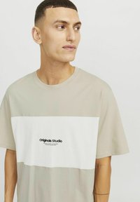 Light beige short-sleeve t-shirt with a white horizontal stripe, featuring a "Originals Studio" logo in black at the center. Smooth texture.