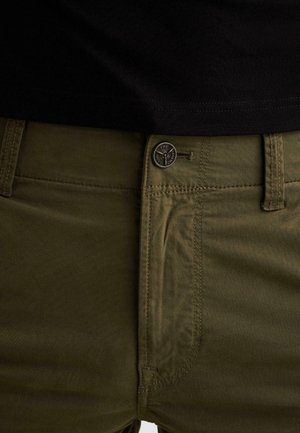 Close-up of olive green pants waistband with button closure and topstitching, worn with a black shirt.