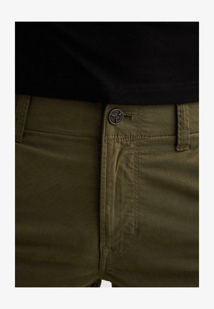 Približeni prikaz pojasa maslinasto zelenih hlača s kopčom i gornjim šavom, nošenih s crnom košuljom.