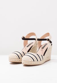 Espadrillas a zeppa a righe con tacco in juta intrecciata, tomaia in tessuto a righe nere e beige, e cinturino alla caviglia con hardware in tono oro.