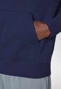 Felpa blu navy con tasca a marsupio, polsini a costine e una texture morbida, abbinata a pantaloni sportivi grigio chiaro.