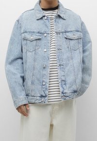 Personne portant une veste en jean bleu clair par-dessus une chemise rayée noir et blanc et un pantalon blanc cassé, debout devant un fond blanc.
