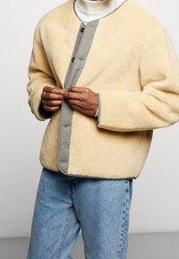 Giacca di colore crema foderata in pile con una placca dei bottoni grigia, design oversize e texture morbida, abbinata a jeans in denim azzurro chiaro.