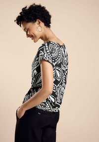 Top a maniche corte bianco e nero, con una vestibilità ampia e scollatura rotonda, abbinato a pantaloni scuri, su uno sfondo neutro.