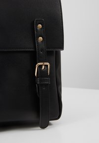 Sac en cuir noir avec une boucle dorée et deux rivets dorés sur la sangle avant, présenté sur un fond gris.
