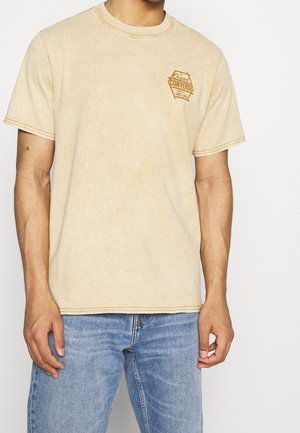 Beżowa koszulka z krótkim rękawem z małym brązowym logo Converse na lewej piersi, noszona przez osobę w niebieskich dżinsach na jednolitym tle.