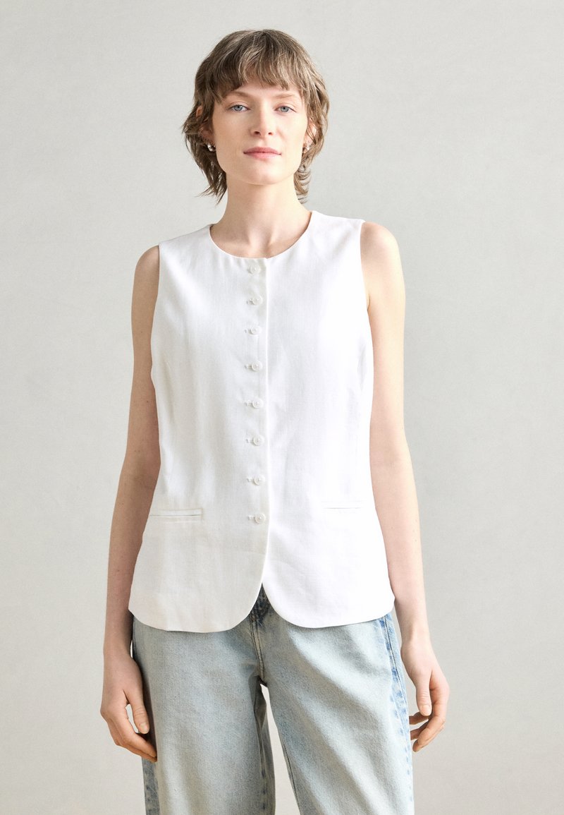 Blouse sans manches en lin blanc avec un col rond, une fermeture à boutons sur le devant et deux poches latérales. Texture subtile et design épuré.